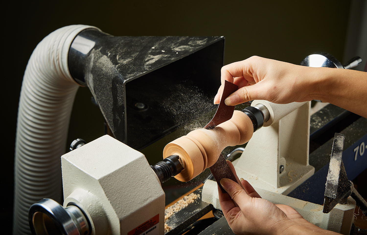 Using a strip of cloth-backed abrasive to sand a turned wooden peppermill as it spins on a lathe.