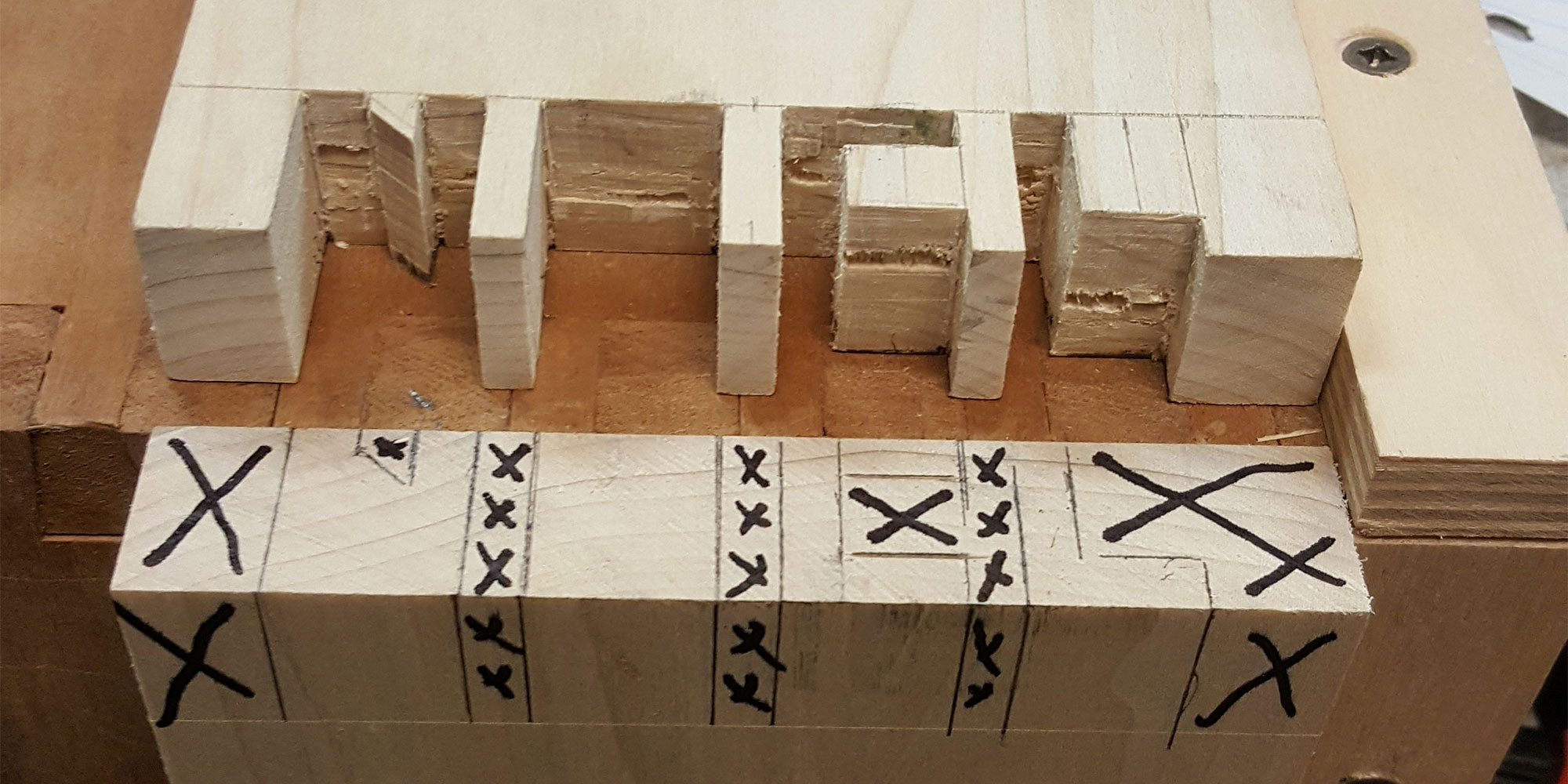 Planche maintenue par un serre-joint avec des portions marquées au feutre devant une planche présentant des lettres découpées