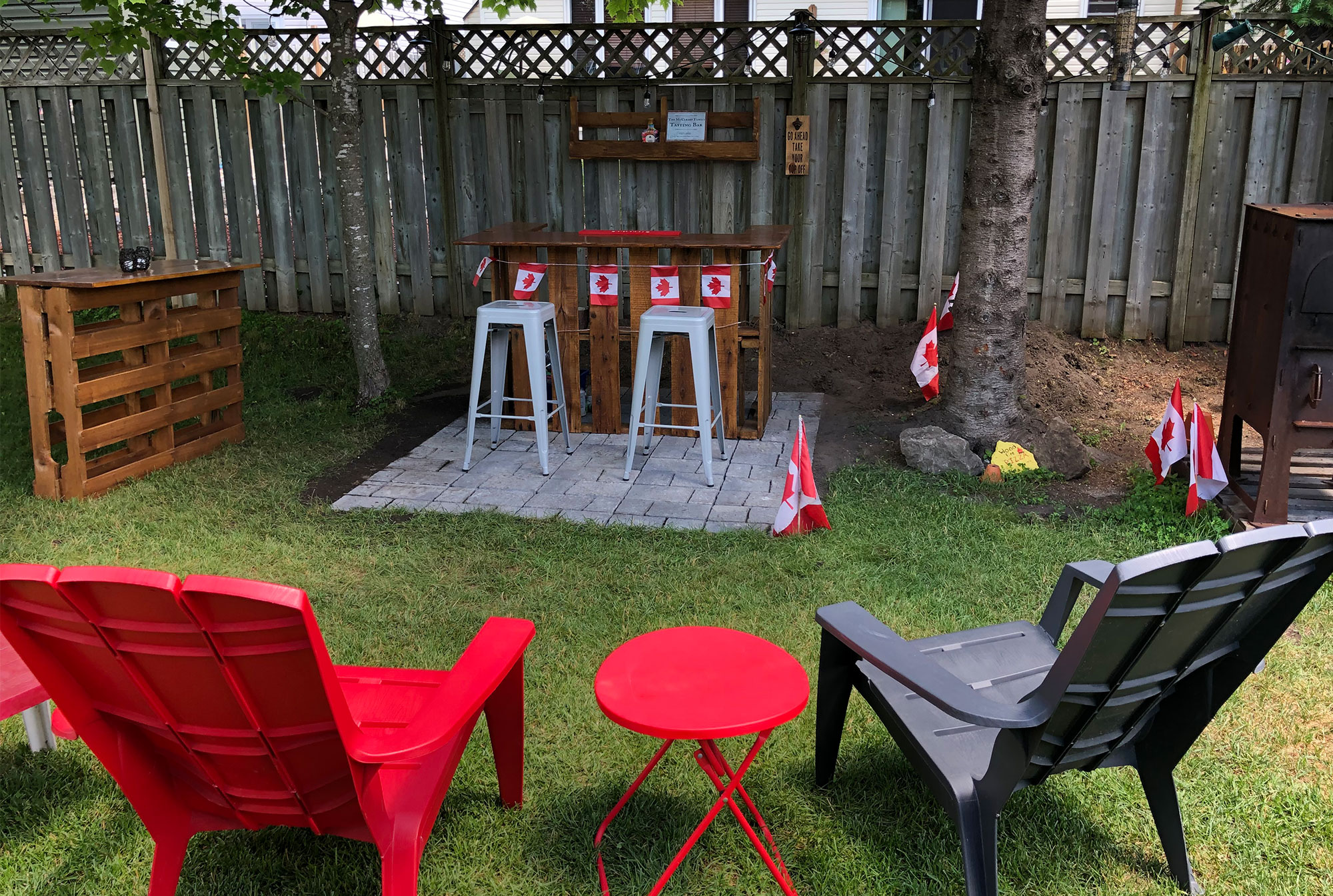 Vue d'une cour arrière avec des chaises de jardin, une table basse, un espace dallé comprenant un bar fait en palettes récupérées, des tabourets et une étagère sur la clôture à l'arrière 